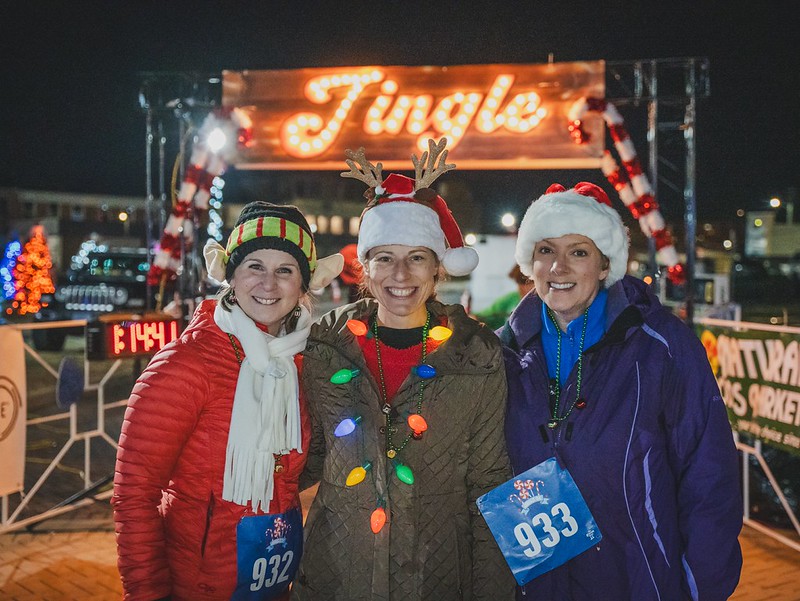 Three smiling participants in holiday hats and race bibs pose beneath the illuminated Jingle sign at a past Jingle event