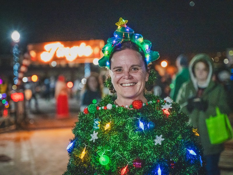 A participant in a lit Christmas tree costume with a star-topped headband smiles in front of an illuminated Jingle sign at a past Jingle event