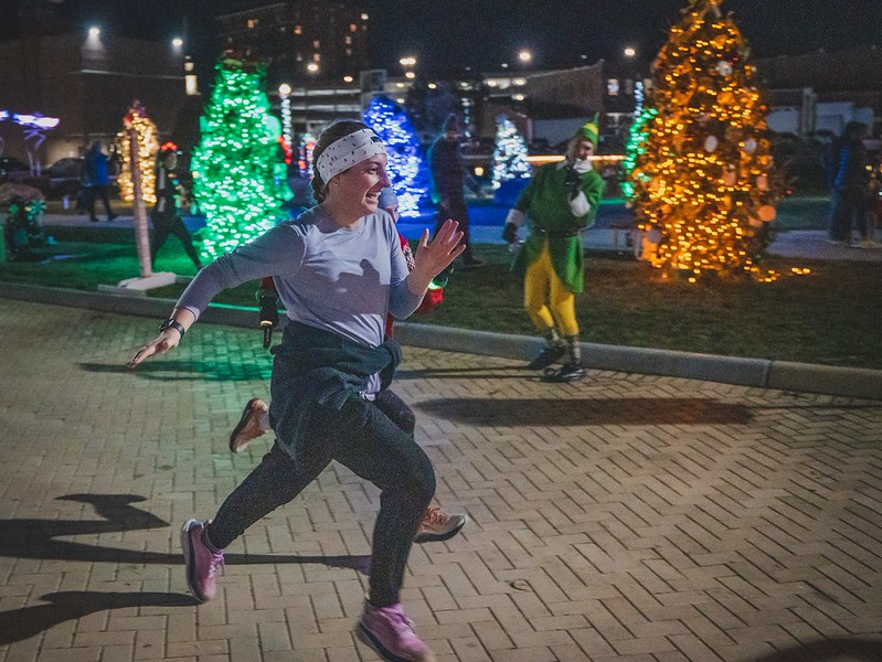 A joyful runner mid-stride smiles as a person in a Buddy the Elf costume cheers her on beside glowing colored trees at a past Jingle event