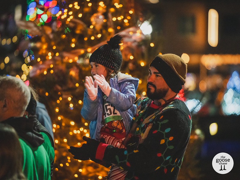 An adult holds a clapping child beside holiday lights at a past Jingle event