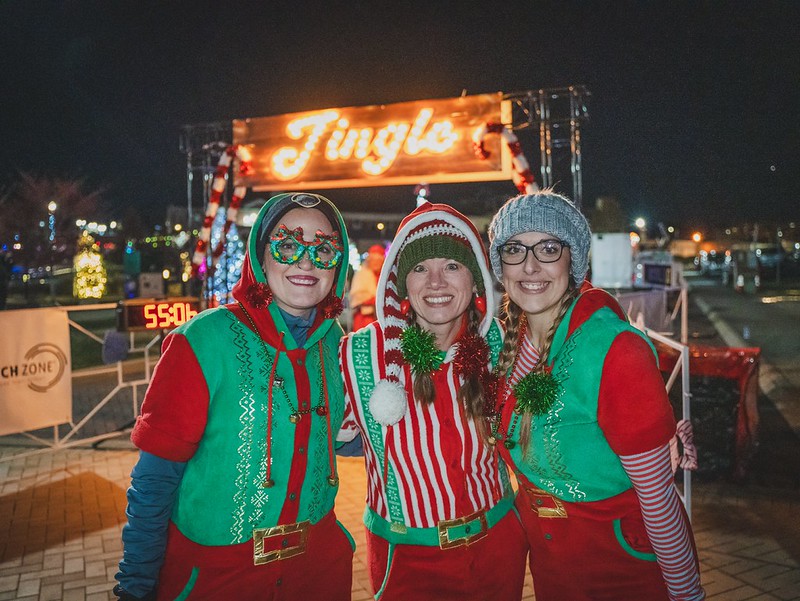 Three participants in green and red elf costumes pose beneath the illuminated Jingle sign at a past Jingle event